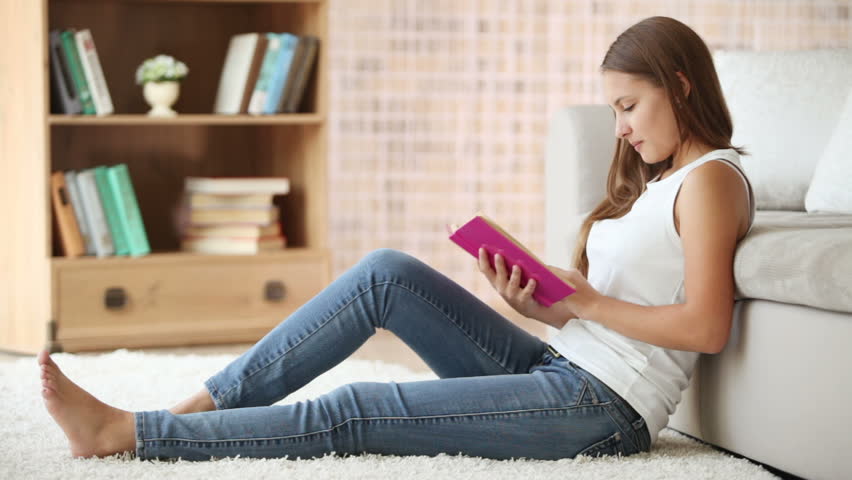 Charming girl sitting on floor reading book and smiling at camera