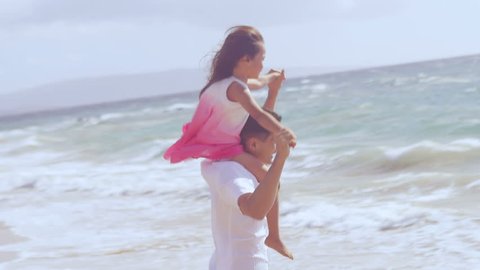 A father holds his daughter on his shoulders and runs around at the edge of the water at the beach