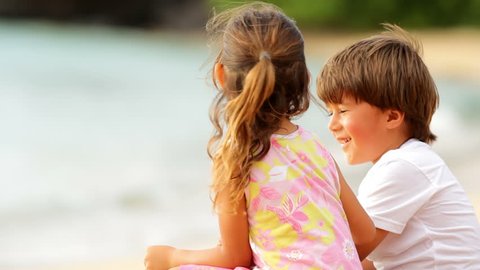 A young boy and girl sit in the sand at the beach and laugh