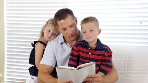 A father sits on a couch reading to his three young children.