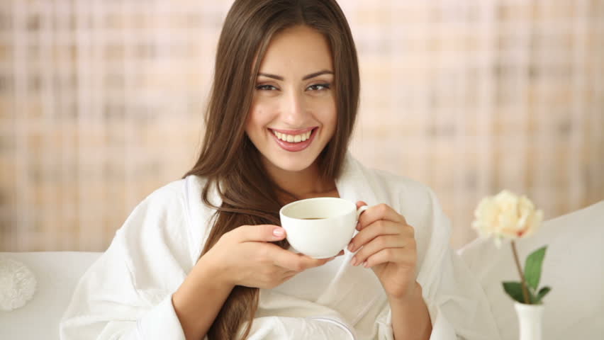 Pretty young woman relaxing in bed drinking tea looking at camera and smiling.