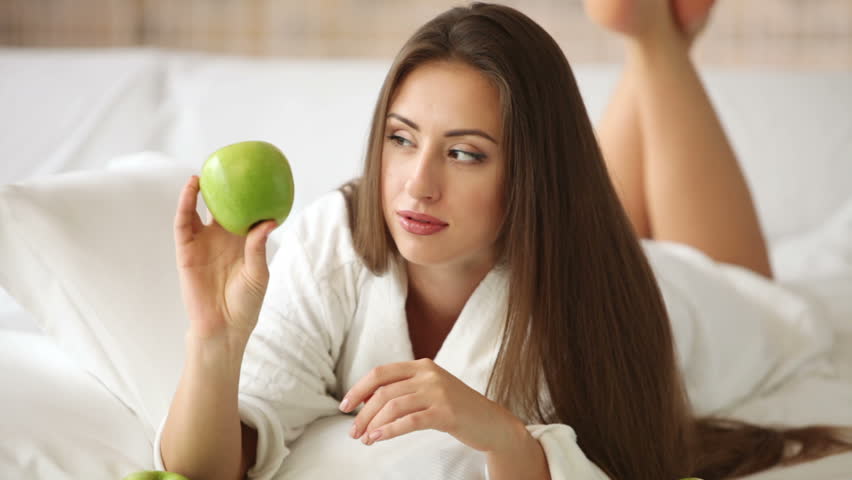 Pretty girl lying on bed with apples in front of her holding one and smiling at