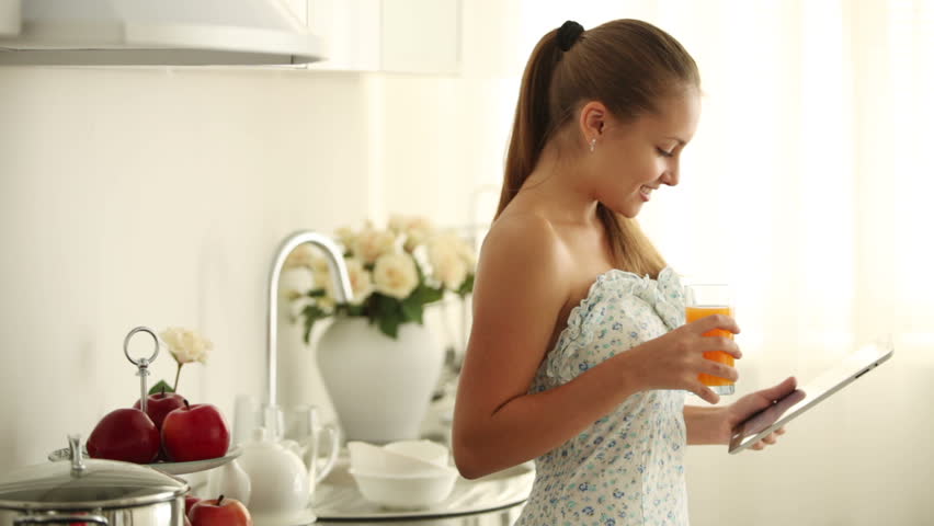 Cheerful girl standing in kitchen drinking juice holding touchpad and smiling at