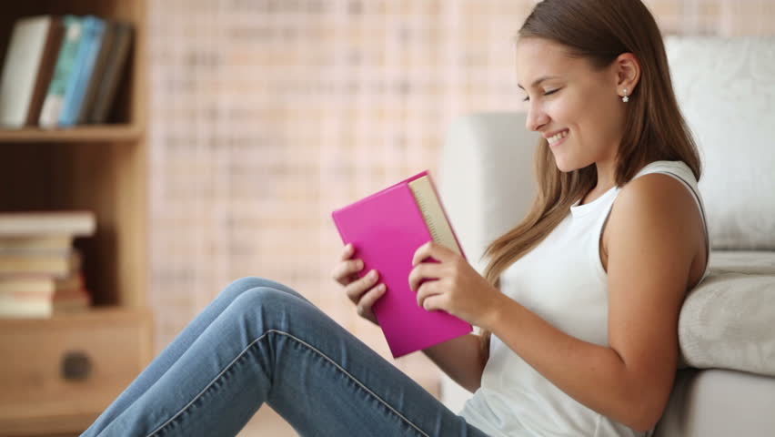 Cute girl sitting on floor reading book closing it and smiling. Panning camera
