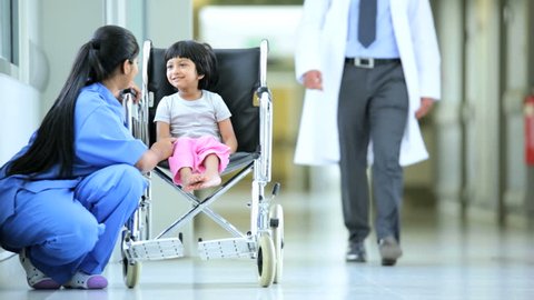 South Asian nurse and pediatrician reassure cute little girl in wheelchair in hospital corridor