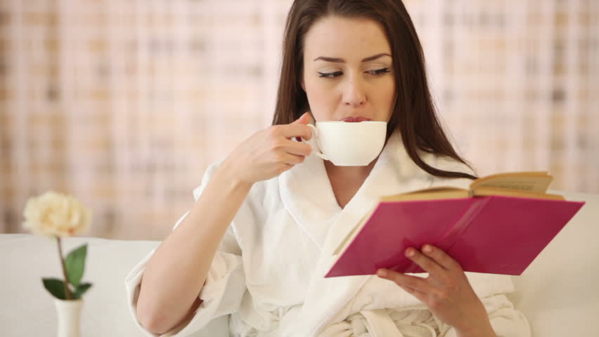 Pretty girl sitting in bed drinking tea reading book and smiling at camera.