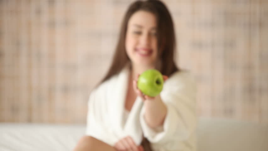Cute girl sitting on bed holding apple showing it at camera smiling and