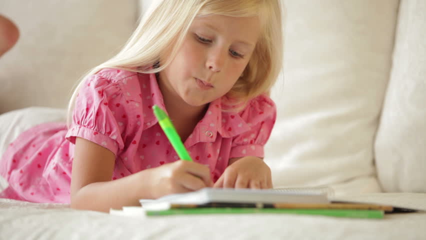 Cheerful little girl lying on sofa writing in notebook and smiling