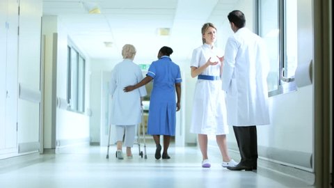 Multi-ethnic medical professionals talking in hospital corridor while elderly patient tries walking for first time after surgery