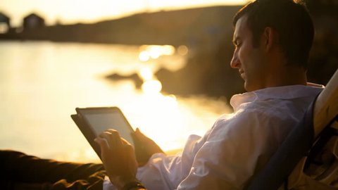 A man looks at his tablet as he sits overlooking the ocean at dusk