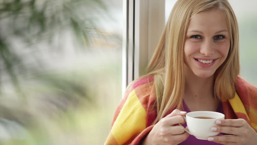 Cute girl sitting by window drinking tea looking at camera and smiling. Panning