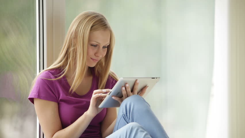 Cute girl sitting by window using touchpad looking at camera and smiling.
