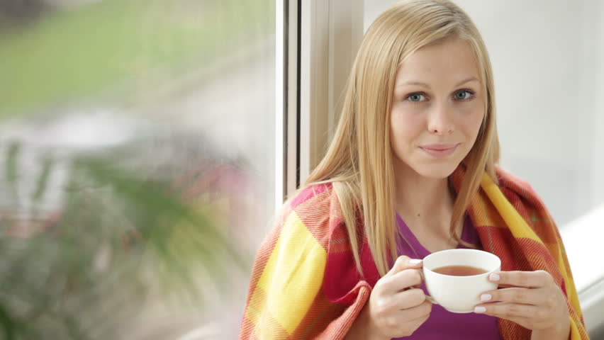 Cute girl wrapped in plaid blanket sitting by window drinking tea looking at