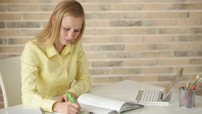 Cheerful student girl sitting at table writing in notebook looking at camera and