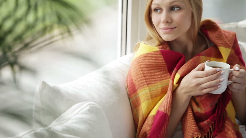 Cheerful girl relaxing on sofa by window drinking tea looking at camera and