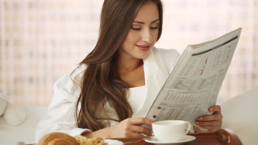Cute young woman relaxing in bed drinking tea and reading newspaper. Panning