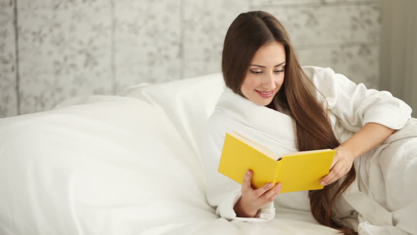 Cute girl in bathrobe lying in bed reading book closing it and smiling at