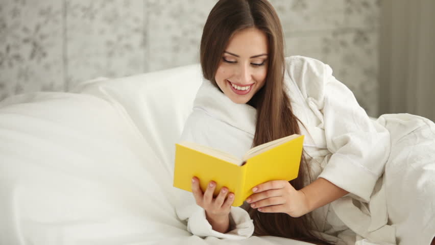 Pretty girl in bathrobe lying in bed reading book looking at camera and smiling.