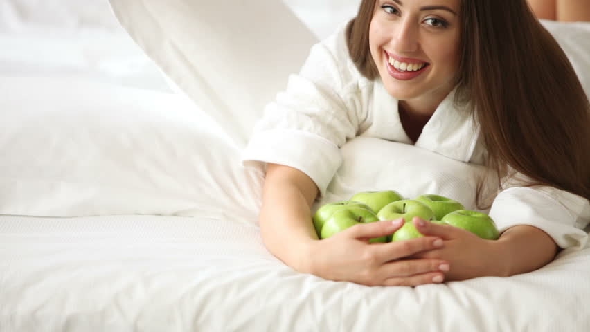 Cute girl lying on bed with lot of apples smiling and laughing at camera.