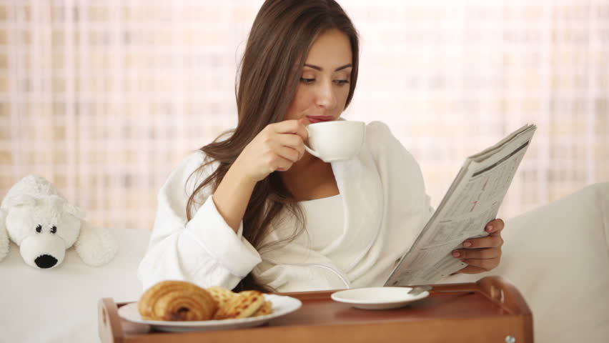 Cute young woman relaxing in bed reading newspaper drinking tea and smiling.