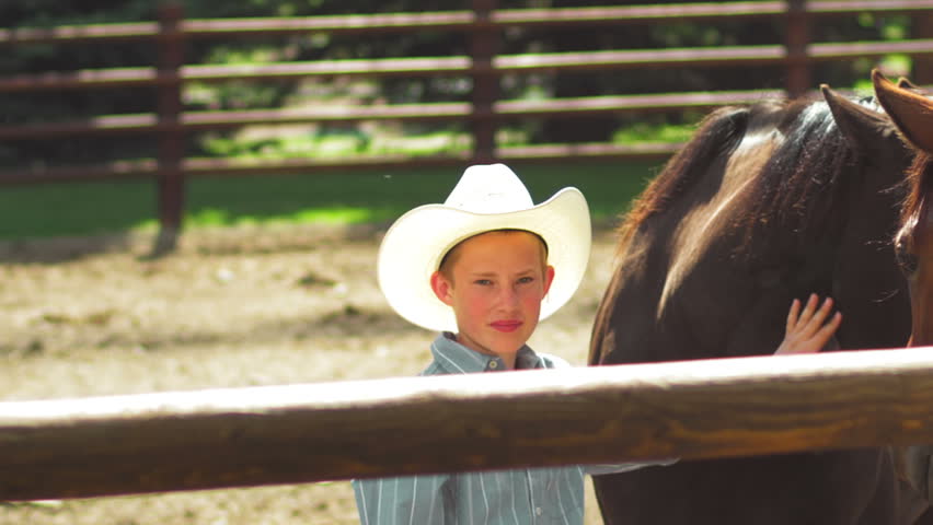 Close up of a boy wearing a cowboy hat standing in a pen and petting a horse