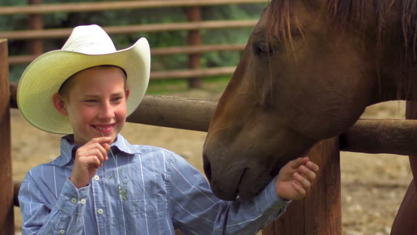 A boy wearing a cowboy hat and chewing straw, stands next to a horse and pets his face