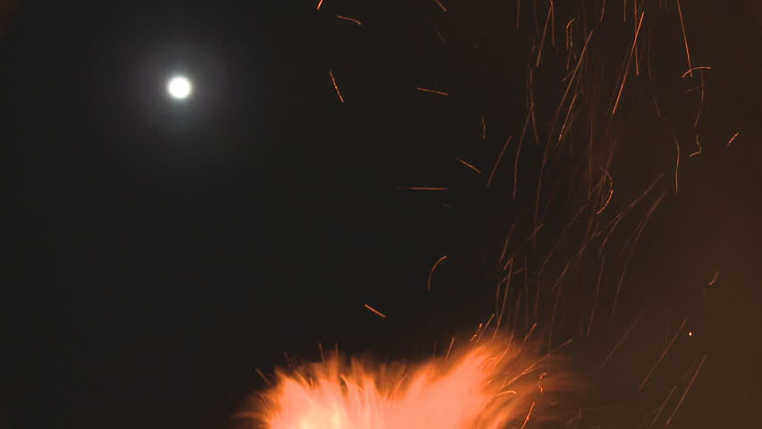 A night time fire sends flames into the air with a full moon visible in the