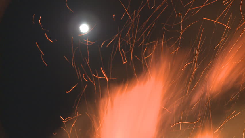 A night time fire sends flames into the air with a full moon visible in the