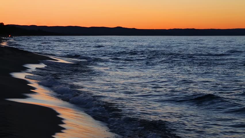 Loop features waves splashing on the Michigan coast of Lake Superior with a colorful sunset sky reflected upon wet beach sand.