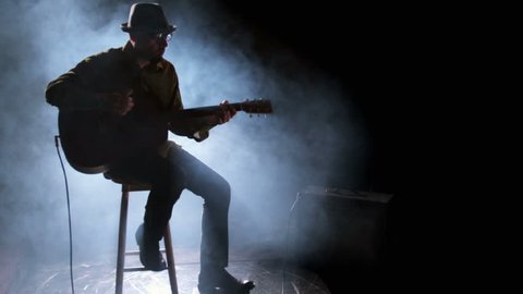 A man sits on a stool playing guitar, backlit in a dark room with smoke blowing behind him  Stock Video