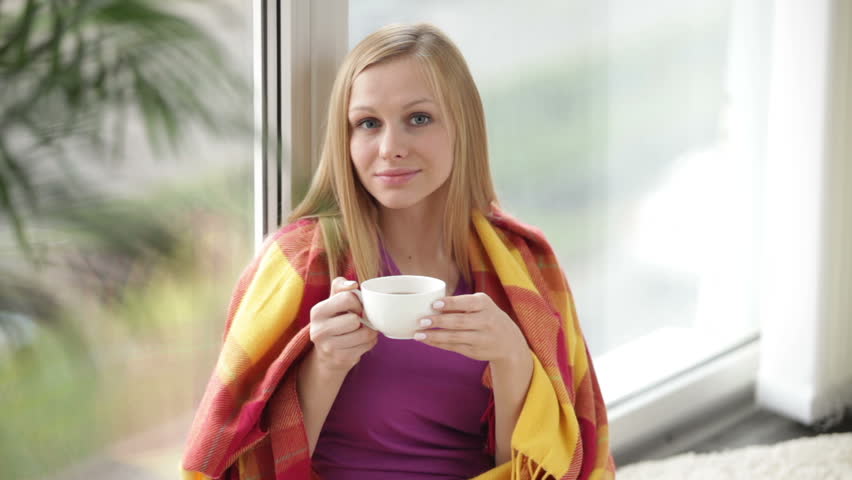 Attractive girl sitting by window drinking tea looking at camera and smiling.