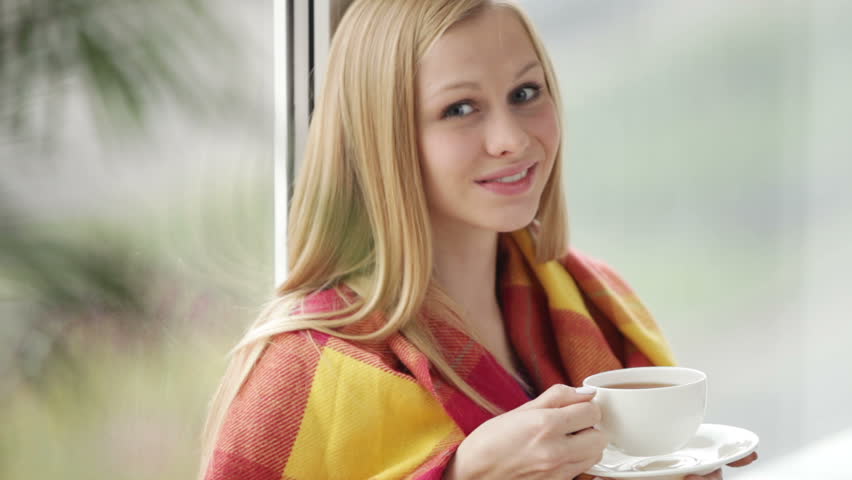 Beautiful girl sitting by window drinking tea looking at camera and smiling.