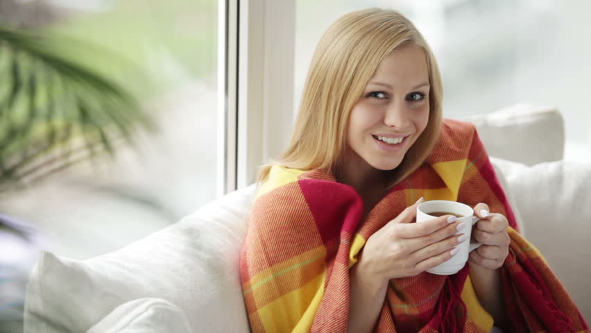 Attractive girl sitting on sofa drinking tea looking at camera and smiling.
