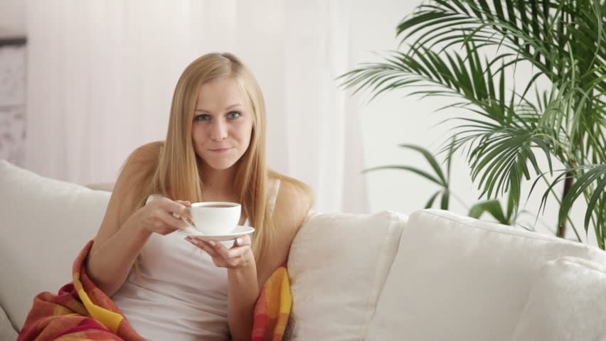 Attractive young woman sitting on sofa drinking tea looking at camera and