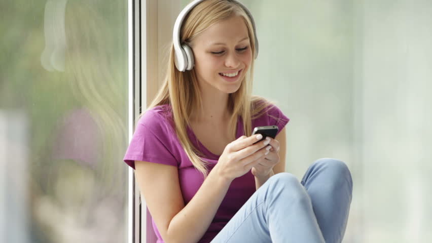Beautiful girl in headphones sitting by window using cellphone looking at camera