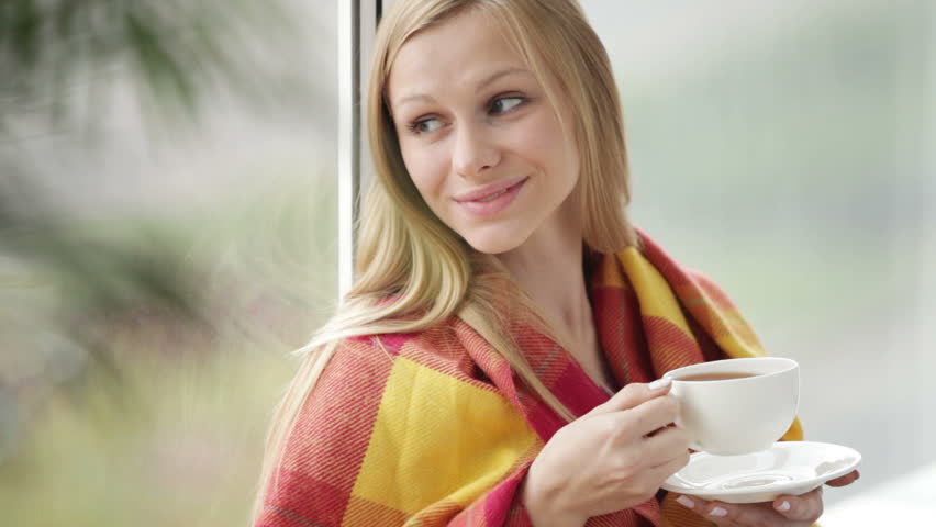Charming girl wrapped in plaid blanket sitting by window drinking tea and