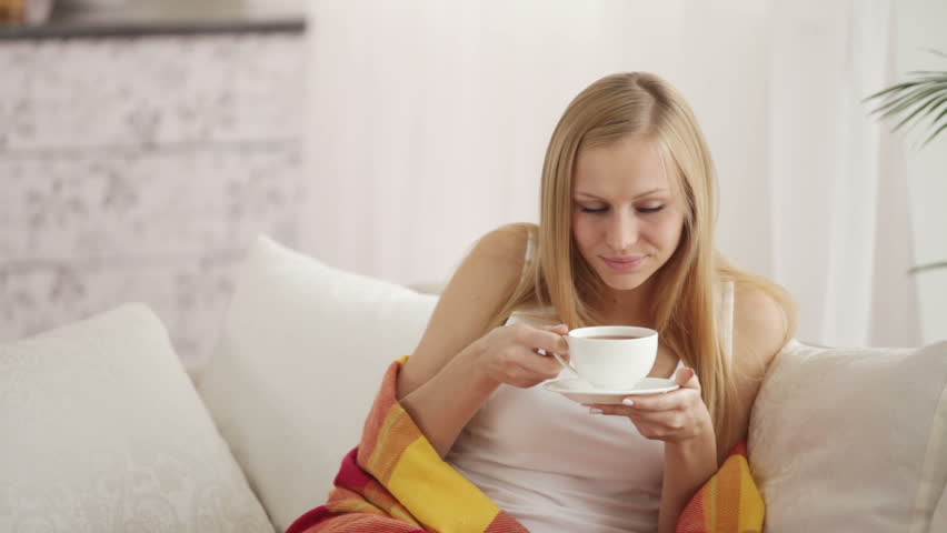 Charming girl sitting on sofa drinking tea looking at camera and smiling.