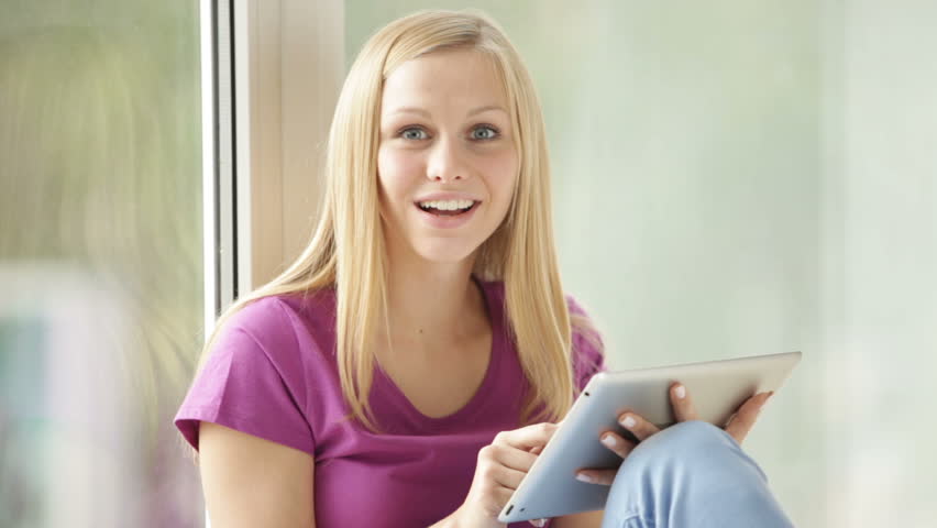 Beautiful girl sitting by window using touchpad looking at camera and smiling.