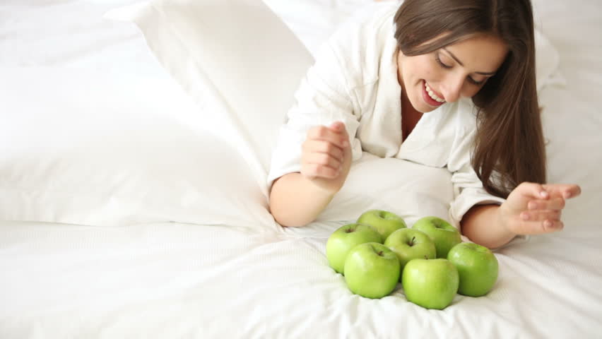 Cheerful young woman lying on bed with lot of green apples looking at camera and