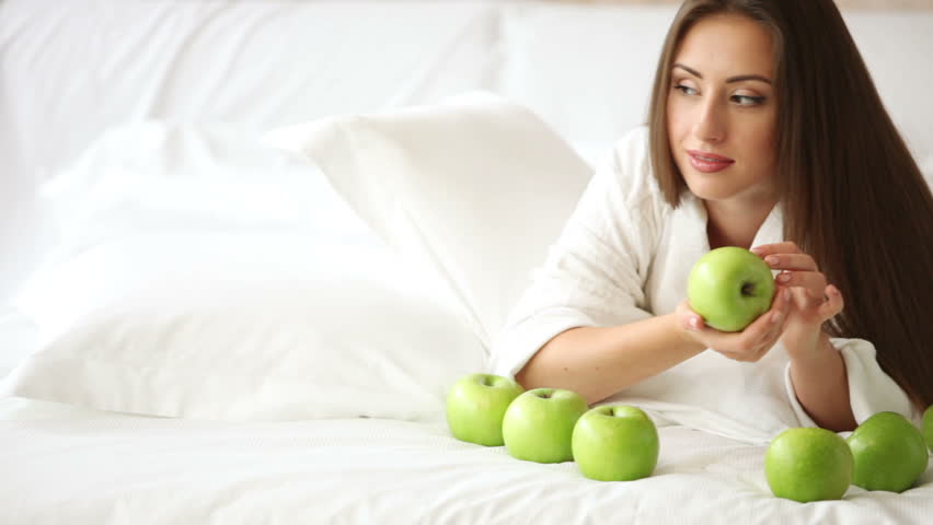 Charming girl lying in bed with apples holding one looking at camera and