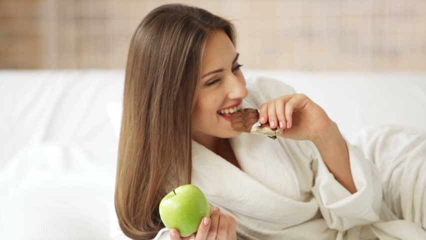 Charming girl lying in bed holding apple and bar of chocolate smiling and