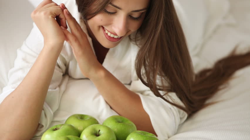 Charming young woman lying on bed with lot of green apples looking at camera and