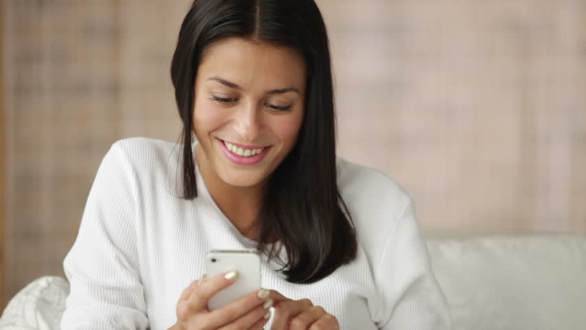 Cute girl sitting on sofa using cellphone looking at camera and smiling. Panning