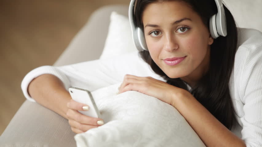Cute girl in headset relaxing on sofa using cellphone looking at camera and
