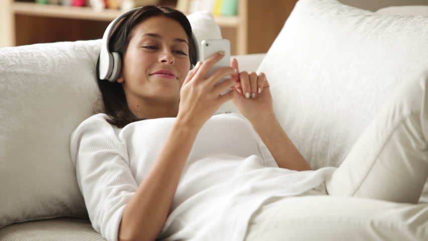 Cute girl in headphones lying on sofa using cellphone looking at camera and