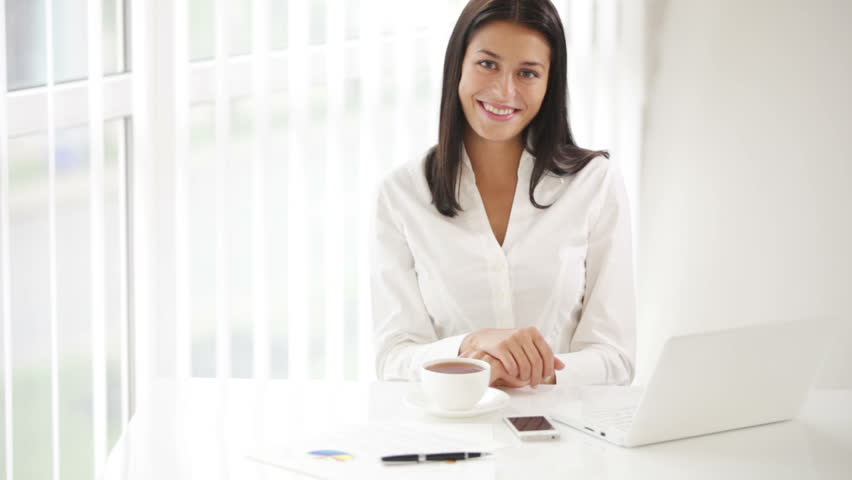 Young woman sitting at office desk with laptop and cup of tea looking at camera