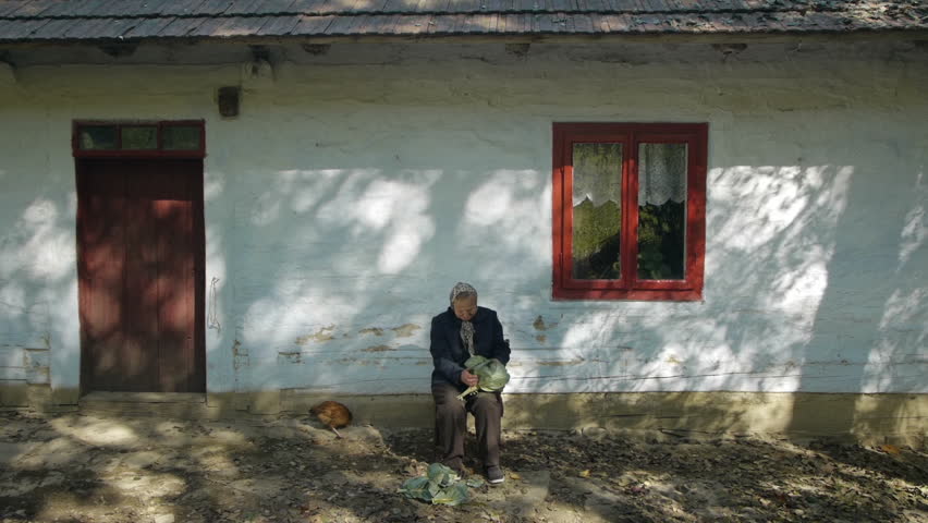 old person peeling lettuce sitting in fornt of a very old white cottage with red window and door