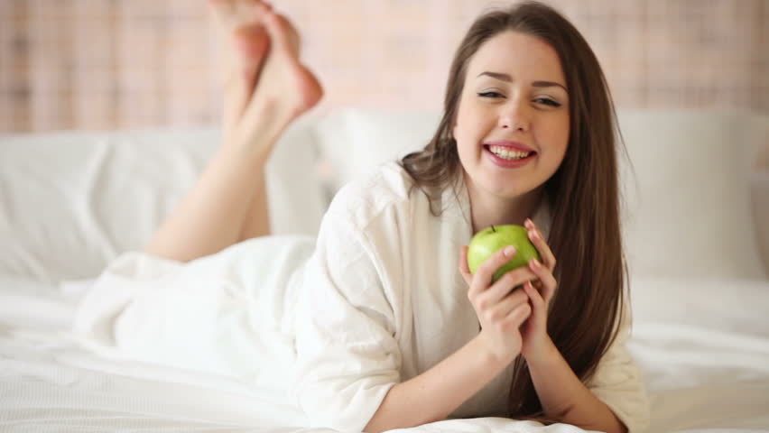 Cute girl lying in bed holding green apple looking at camera and smiling.