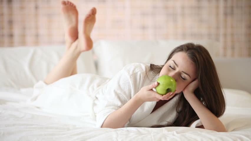 Cheerful girl lying on bed holding green apple looking at camera and smiling