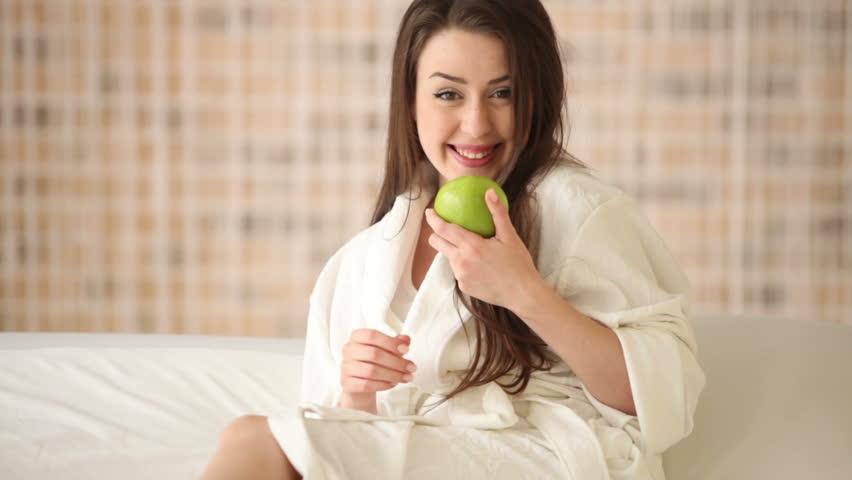 Cheerful girl sitting on bed holding green apple looking at camera and smiling.